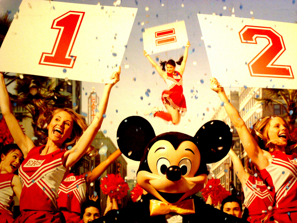A Mickey Mouse mascot flanked by cheerleaders holding signs reading "1" and "2" with an equals sign between them